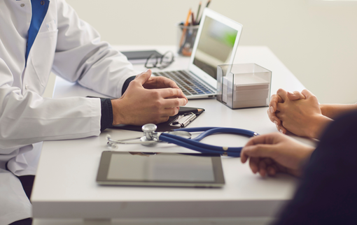Doctor and patient discussing at a clinic desk with a laptop and stethoscope nearby.