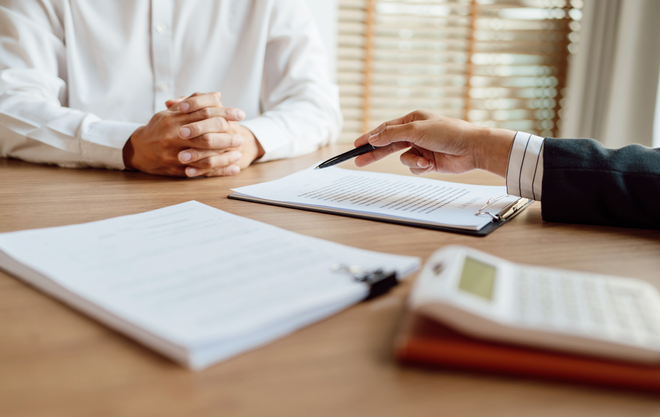Hands discussing paperwork across a desk with a notebook and calculator in a bright office