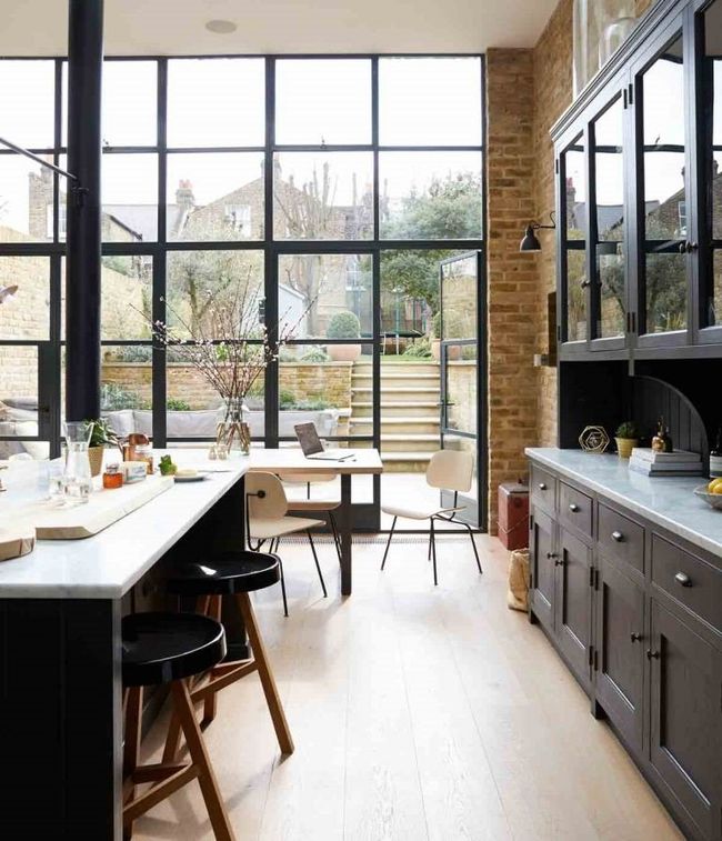 Modern kitchen with black cabinetry, a white island with bar stools, and a large floor-to-ceiling glass wall facing a garden.