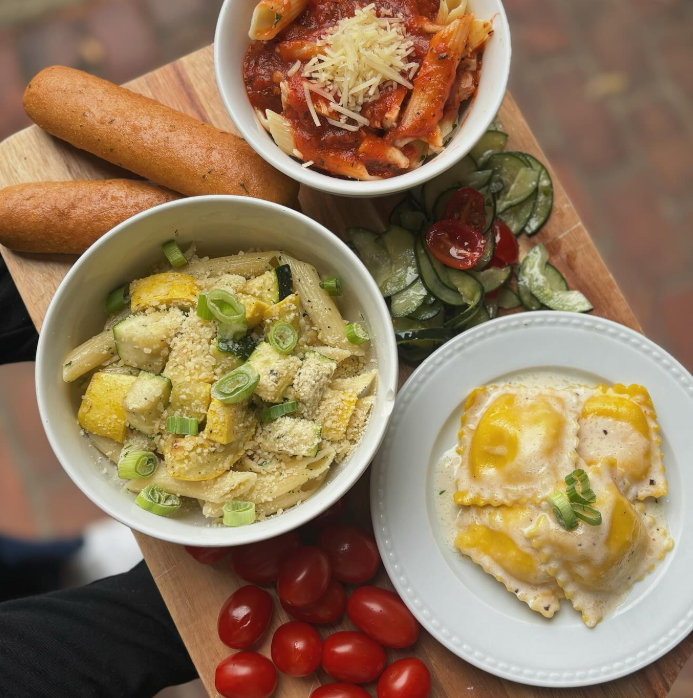Pasta dishes: marinara, pesto, and ravioli with sides on a wooden serving board.