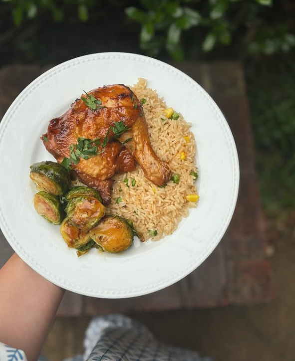 Plate of glazed chicken, fried rice, and Brussels sprouts held in hand, outdoors.