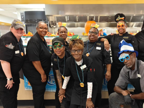 Group of food service workers smiling, posing together in a decorated cafeteria.