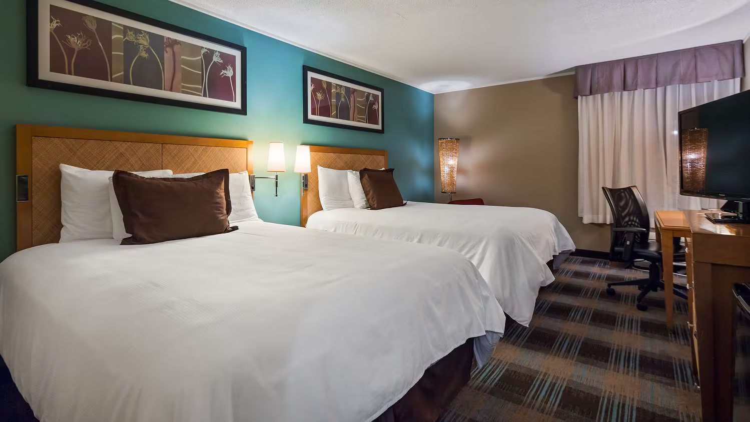 Two double beds in a hotel room with white linens, brown pillows, teal walls, and a desk.