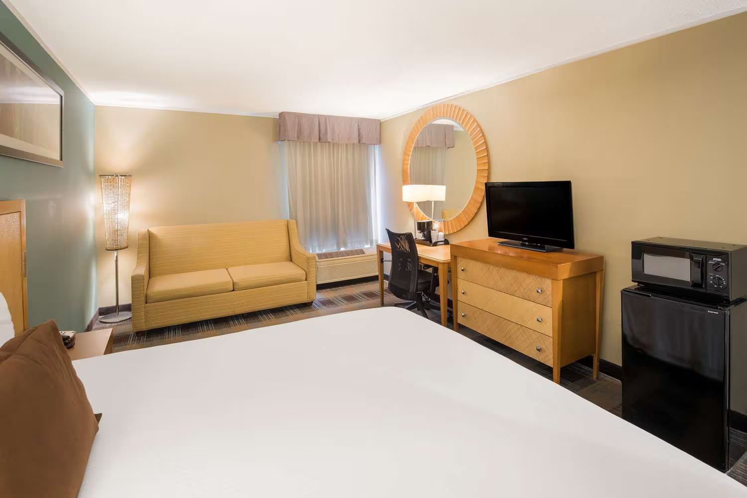 Hotel room with bed, sofa, desk, TV, microwave, and mirror. Beige walls, neutral colors.