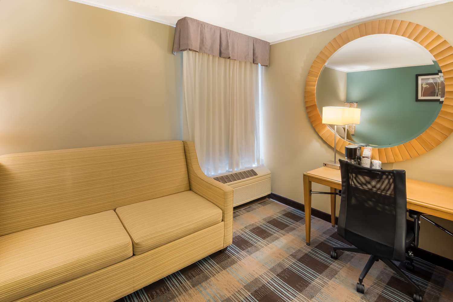 A hotel room with a couch , desk , chair and mirror.