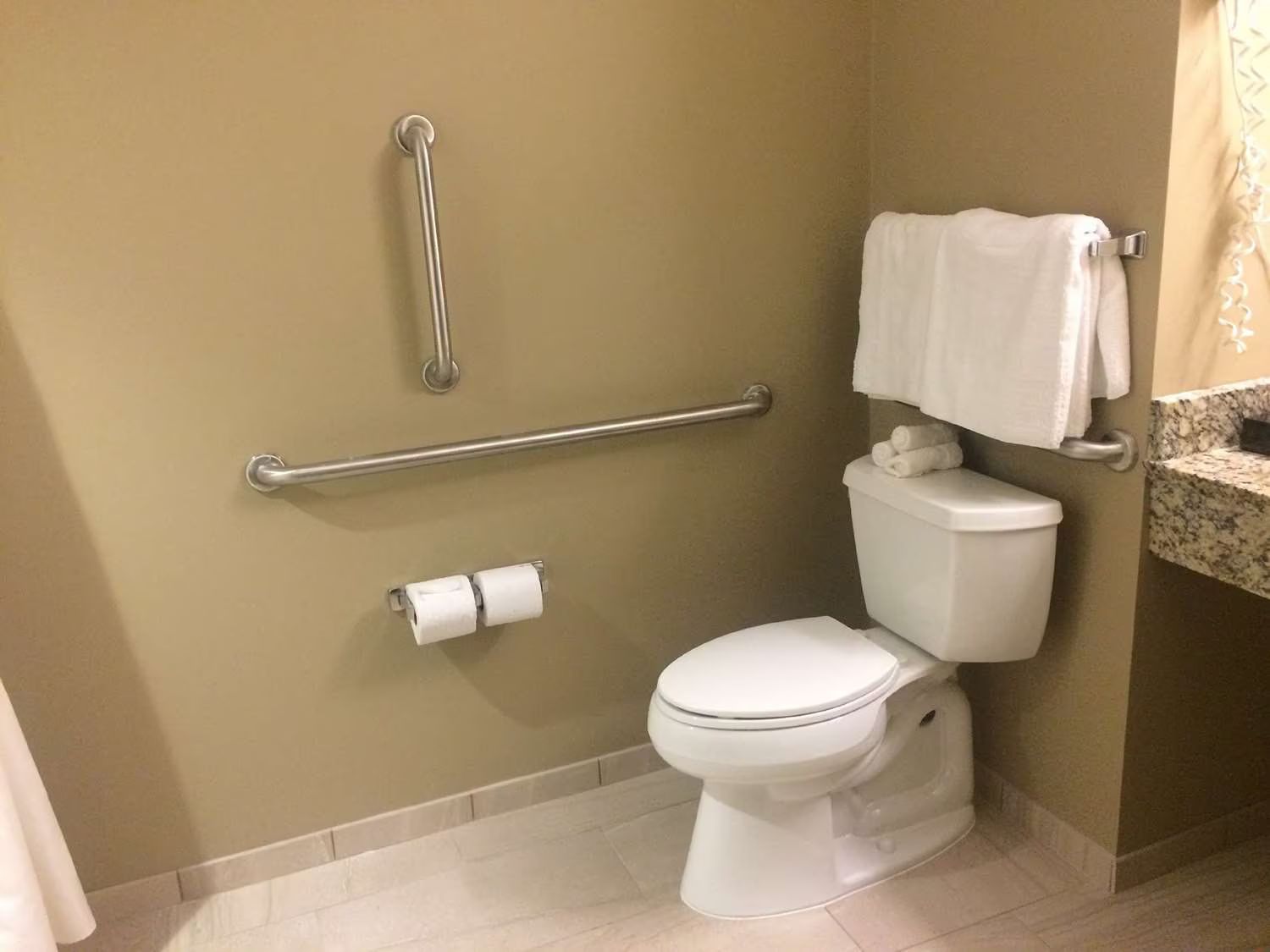 Accessible bathroom with grab bars, toilet, and towel rack. Tan walls, white toilet, and silver fixtures.