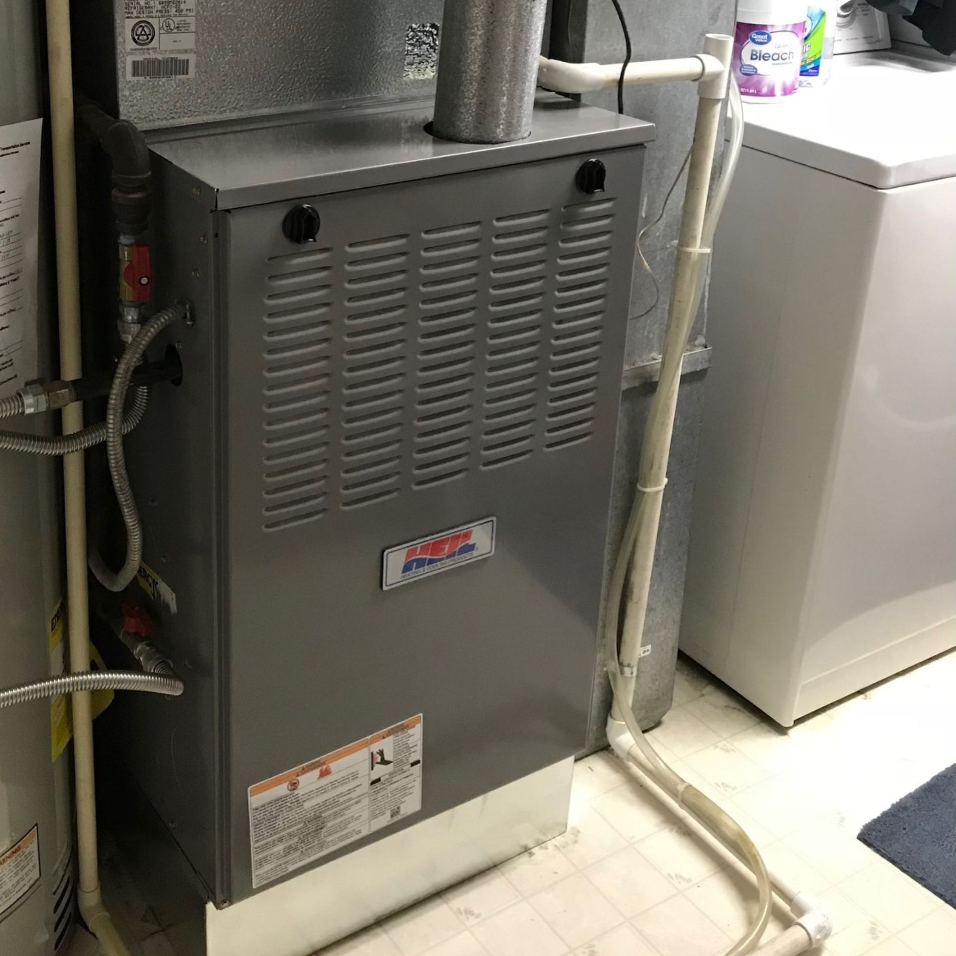 E.Squared Custom Plumbing Heating and Air - A gray furnace in a laundry room with white pipes and a washing machine visible.