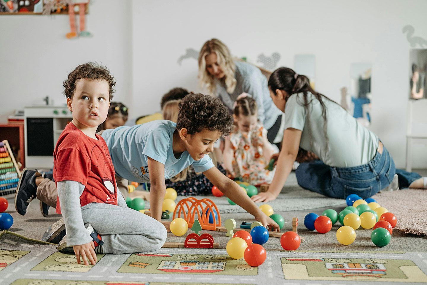 Children playing with colorful balls on rugs in a bright classroom while adults watch nearby