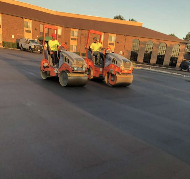 Two men are driving rollers on a road in front of a building