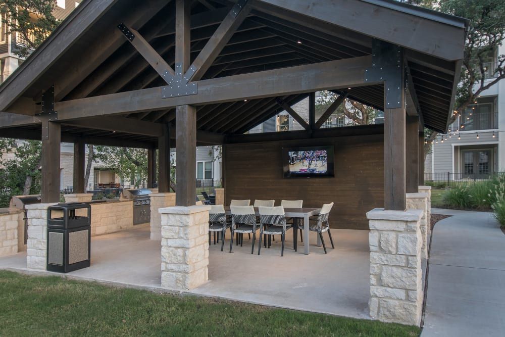 Outdoor pavilion with a dark wood finish and exposed beams supported by metal brackets, featuring a built-in kitchen area with a grill, under a string of lights. In the foreground, a dining area with modern chairs and a table, set on a concrete floor. A flatscreen TV mounted on the back wall displays a sports game, with greenery and apartment buildings visible in the background at Marquis Dominion in San Antonio, TX.