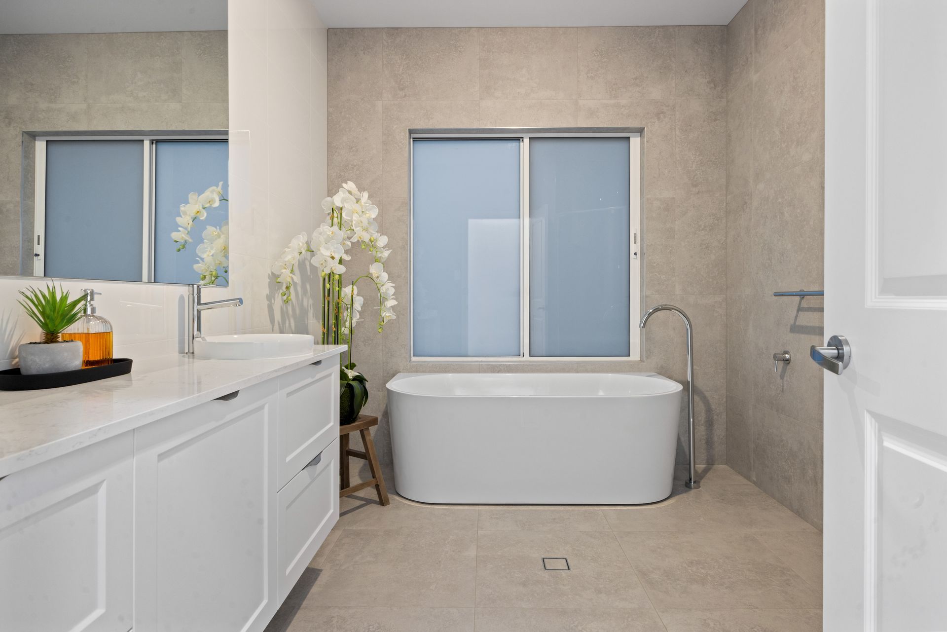 Modern bathroom with white tub, vanity, and neutral tile walls. Window with blue blinds, decorative flowers.