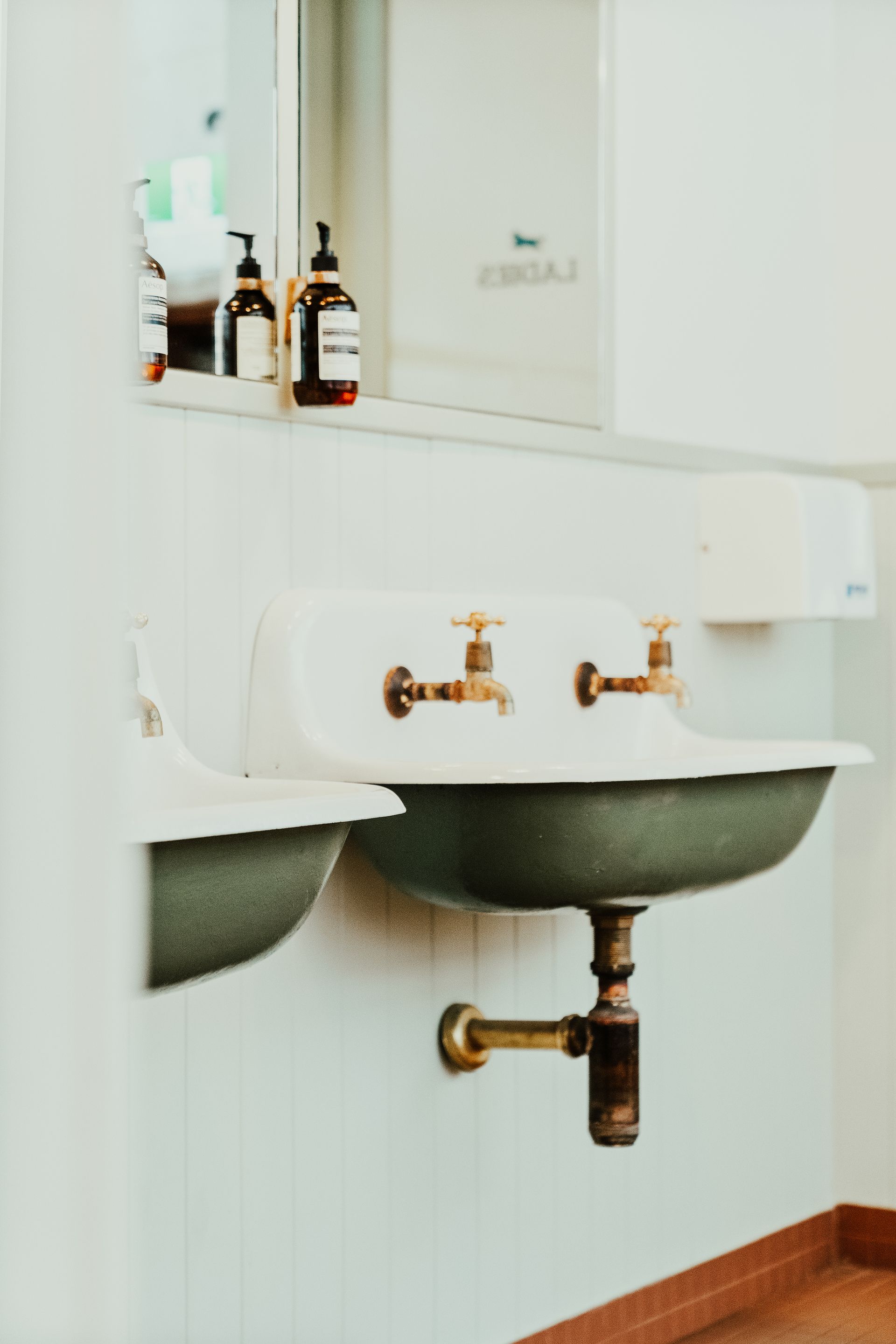 White and green bathroom sinks with brass fixtures under a mirror, bottles on shelf— Croker Plumbing and Gas in Goulburn, NSW