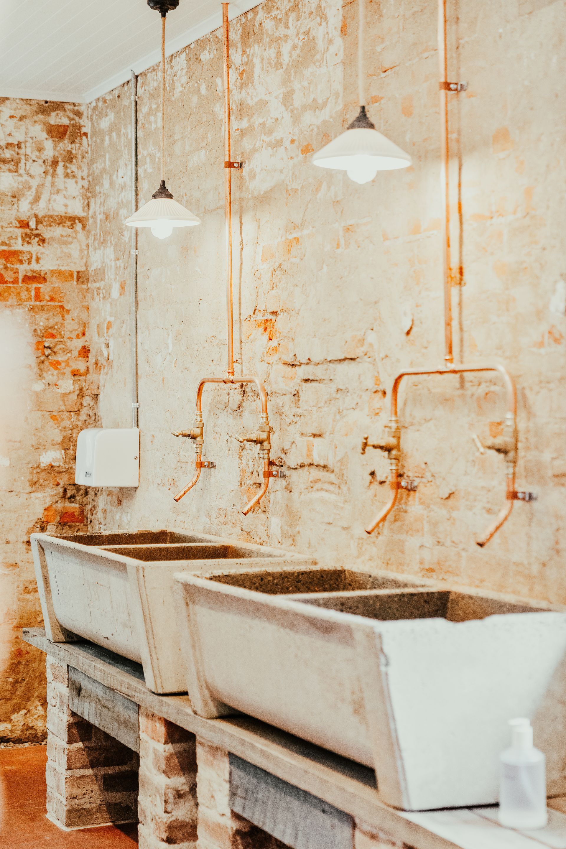 Rustic bathroom with three trough-like sinks, exposed pipes, brick and distressed walls, pendant lights.