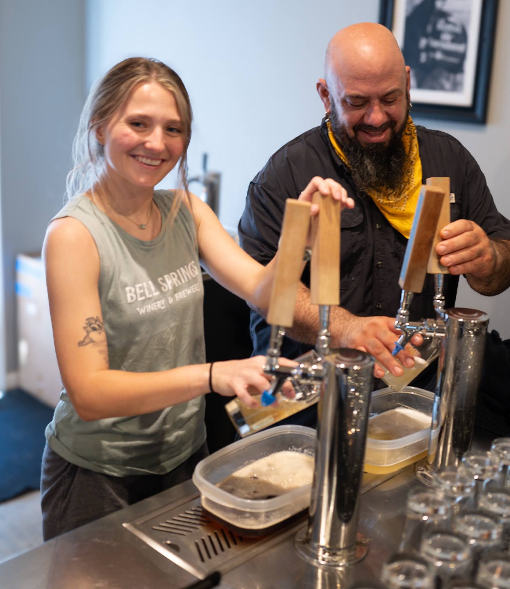 Two people pouring beer from taps behind a bar, smiling.