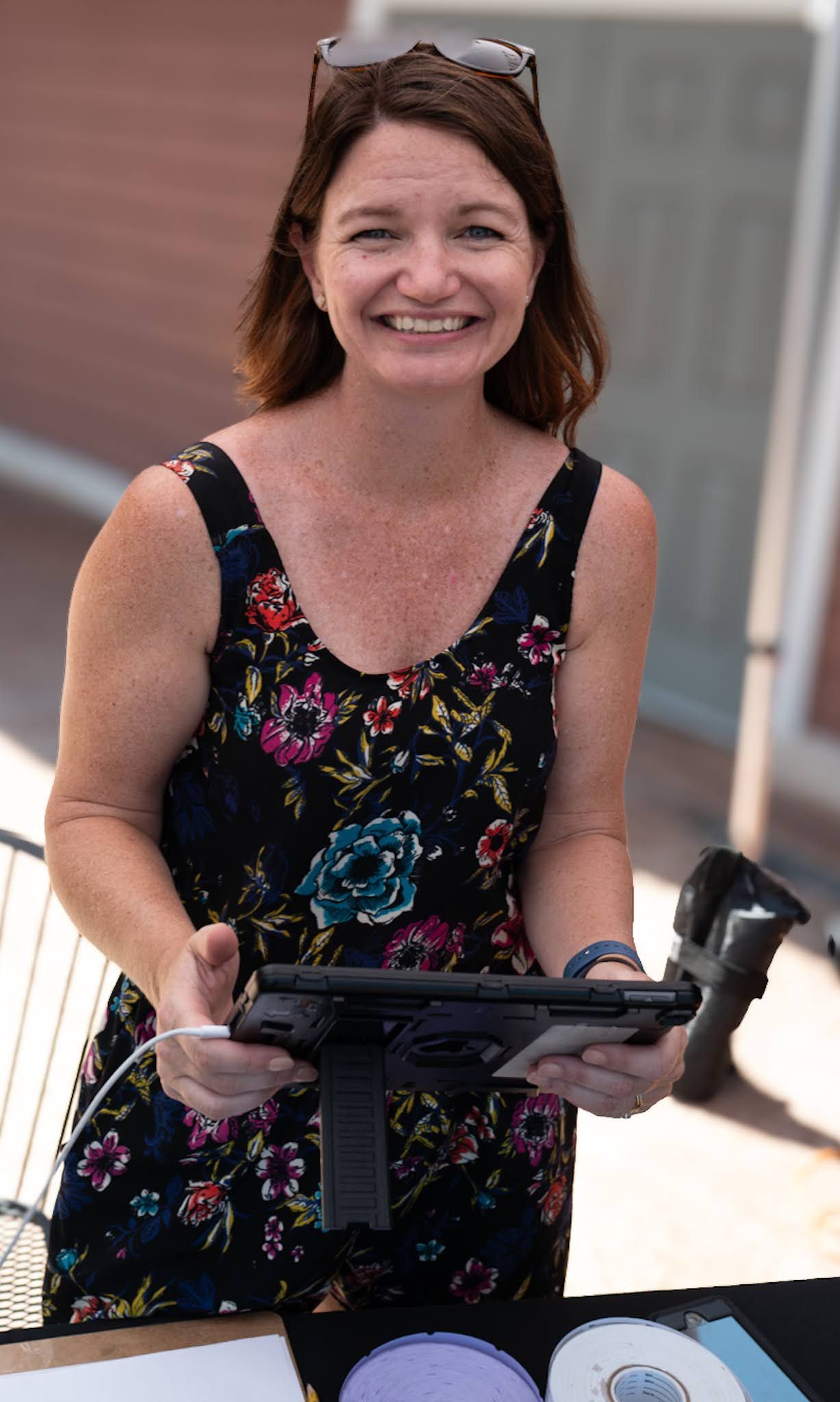Woman smiling, holding a gun. Wearing a floral dress, outdoors. Sunglasses atop her head.