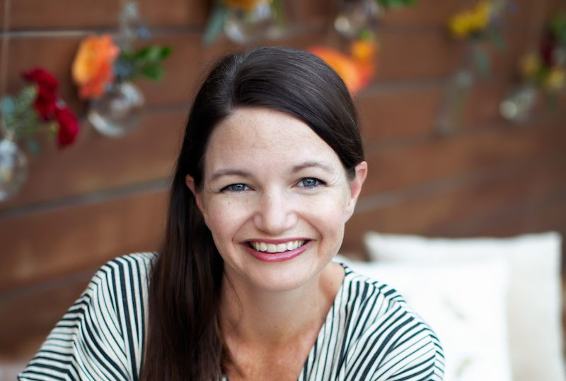 Woman with dark hair smiles, striped shirt, wooden background with hanging flowers.