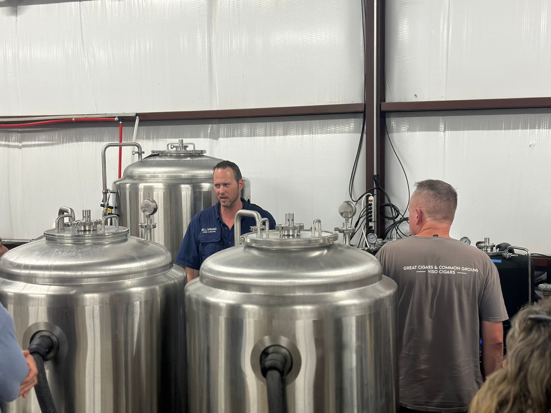 Brewery tour: Man in blue shirt explains equipment. Stainless steel tanks in a warehouse. People listening.