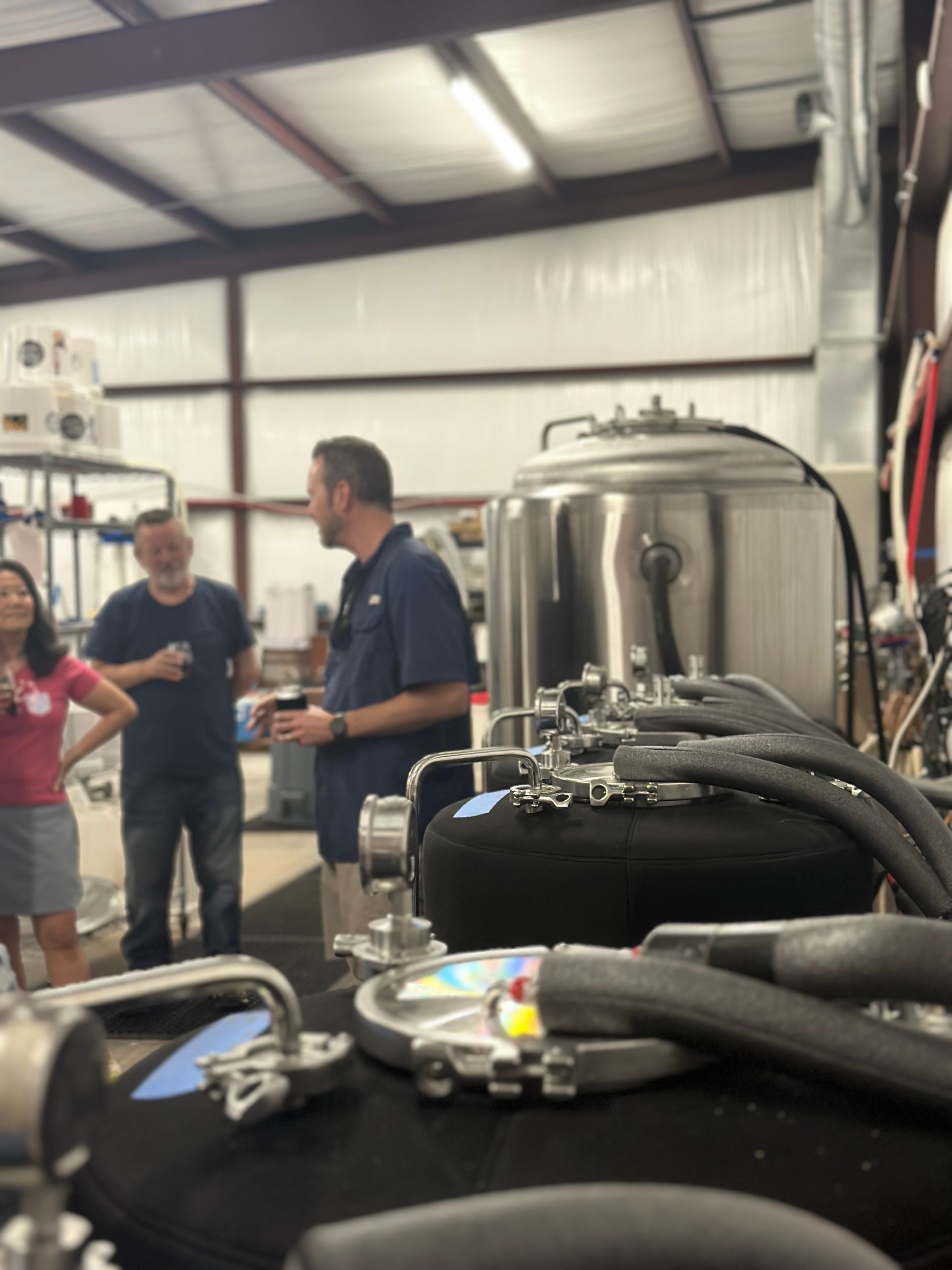 People in a brewery, one man is speaking, looking at a large steel tank and machinery.