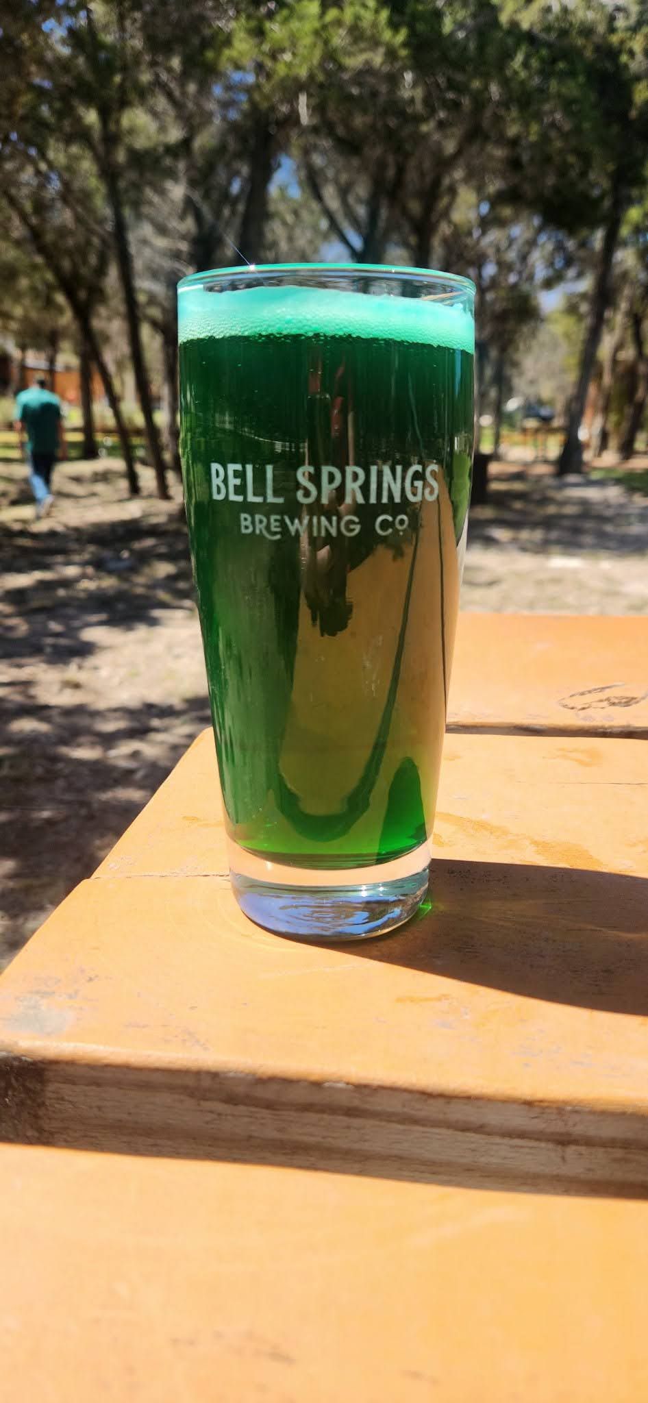 A tall glass of green liquid from Bell Springs Brewing Co. sits on a wooden table in a park.