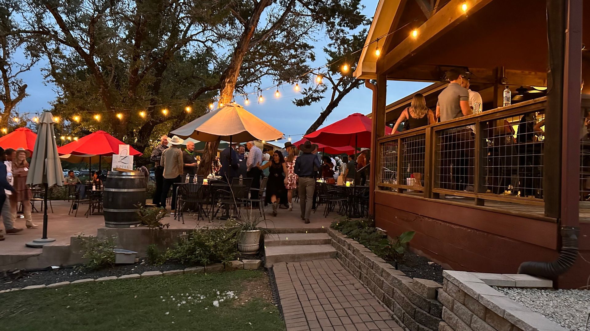 Outdoor evening gathering at a restaurant with people socializing, red umbrellas, and string lights.