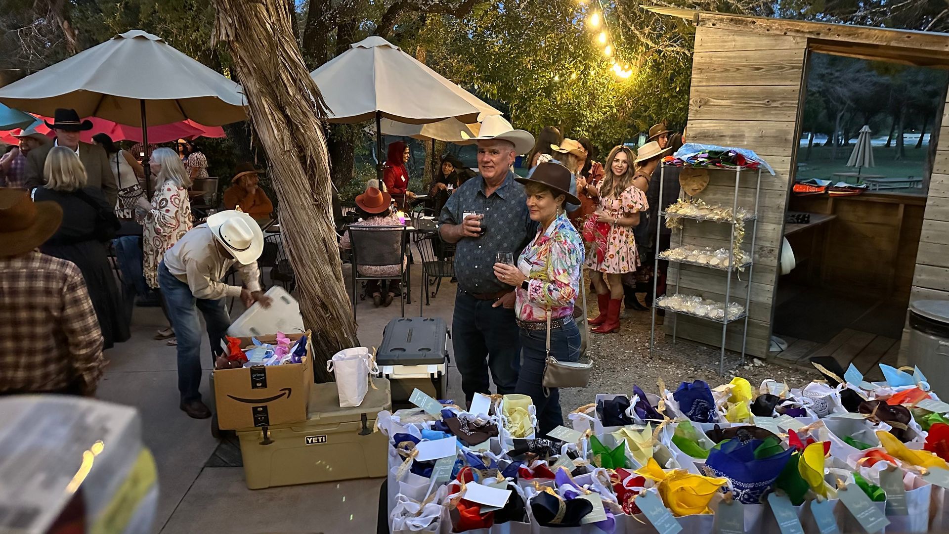 People at an outdoor market, some wearing cowboy hats. Various items on tables, trees and umbrellas overhead.