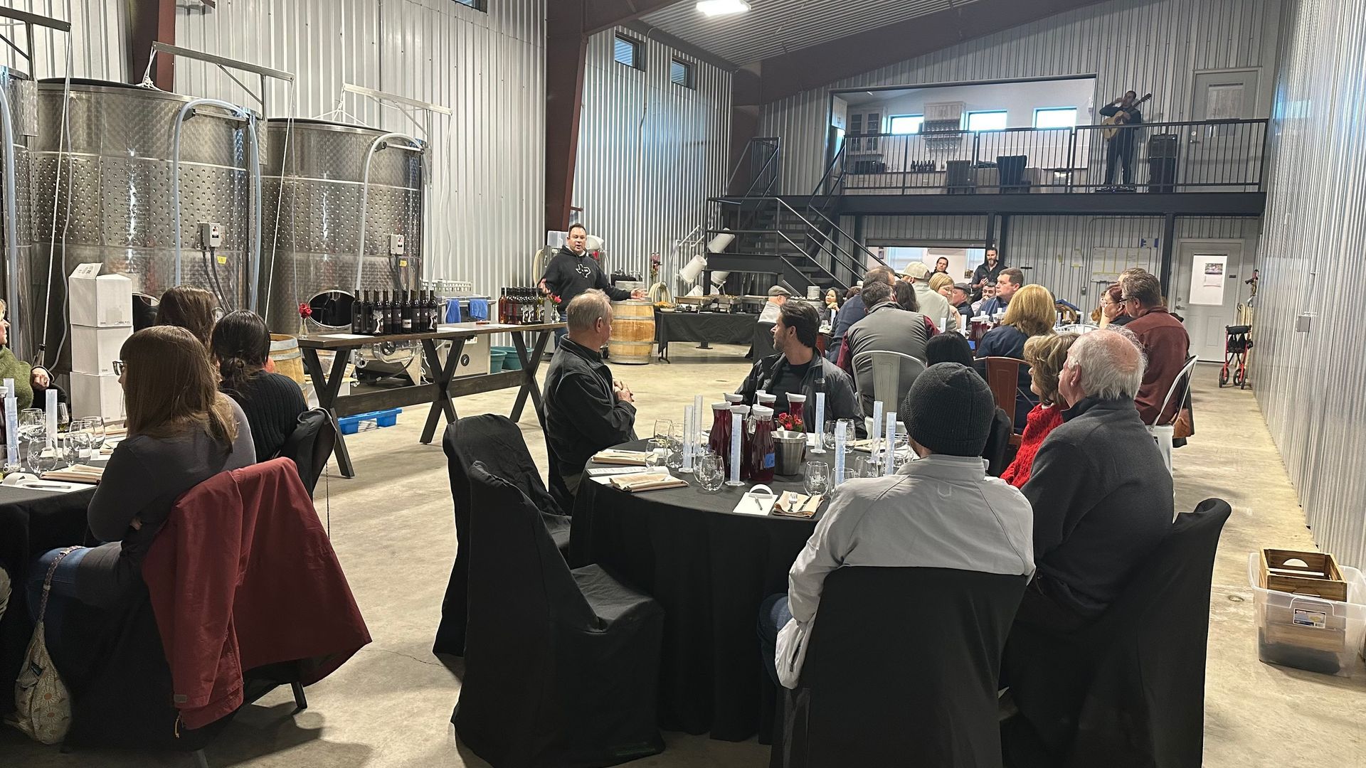 People seated at tables in a winery, attending an event. Stainless steel tanks in the background.