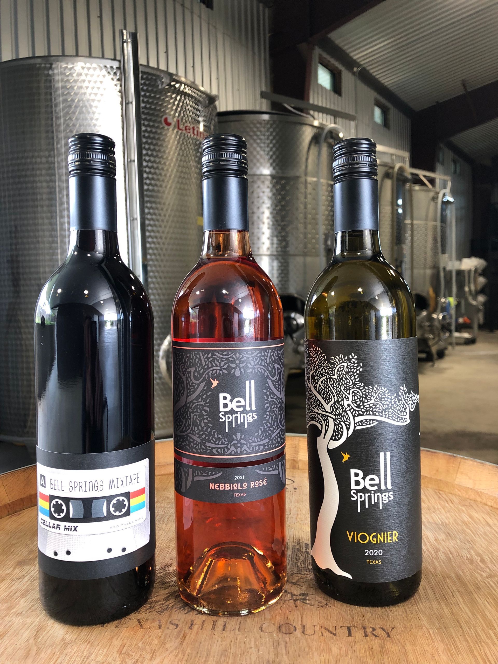 Three bottles of wine: red, rosé, and white, on a wooden surface.  Stainless steel tanks are in the background.