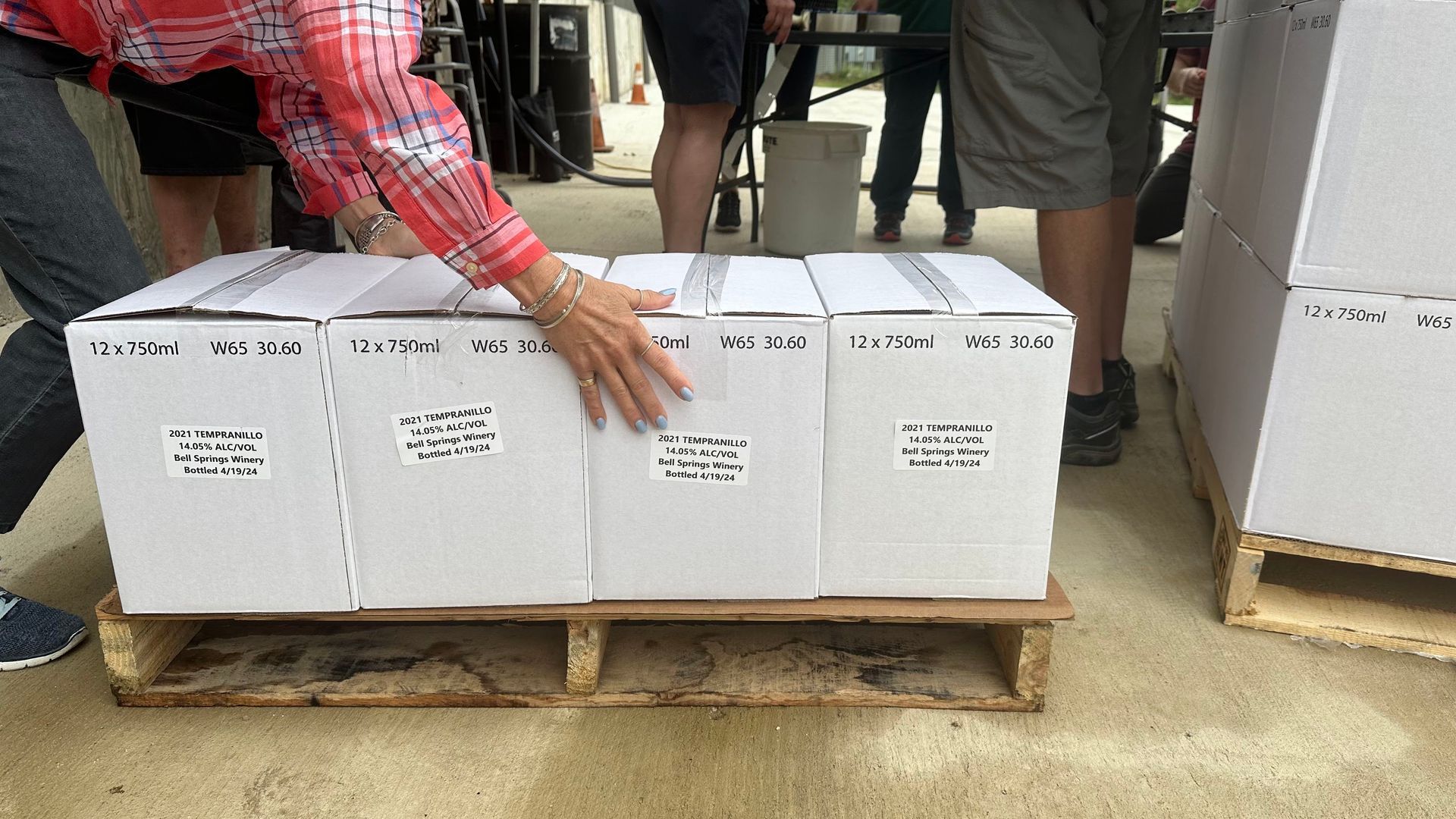 Person arranging white boxes on a wooden pallet. Other people stand nearby.