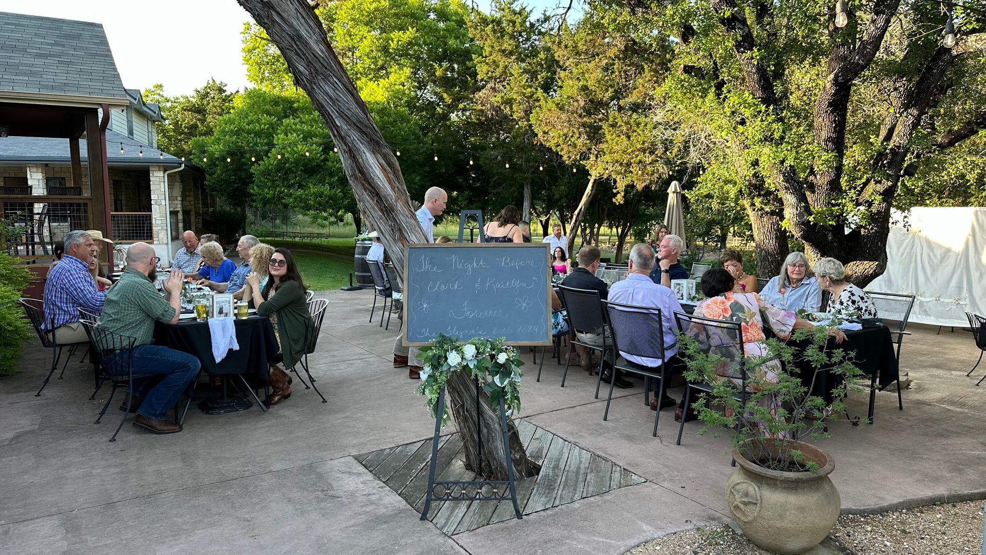 Outdoor gathering: People seated at tables, dining under trees, sign in foreground.