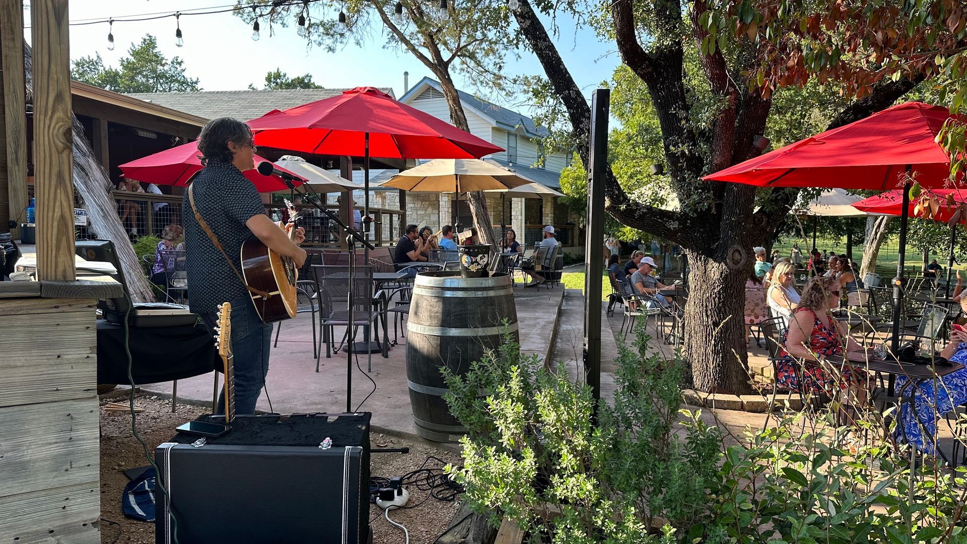 A musician plays guitar outdoors at a restaurant with red umbrellas and a seated audience.
