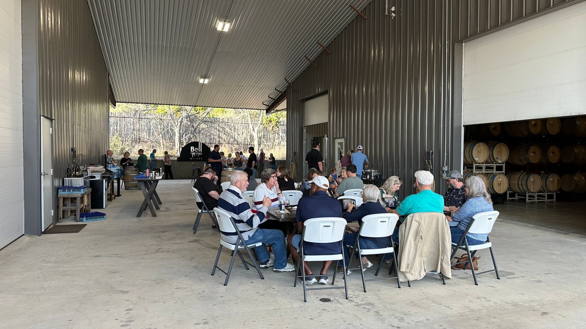 People seated at tables under a metal awning, some drinking, with barrels visible.