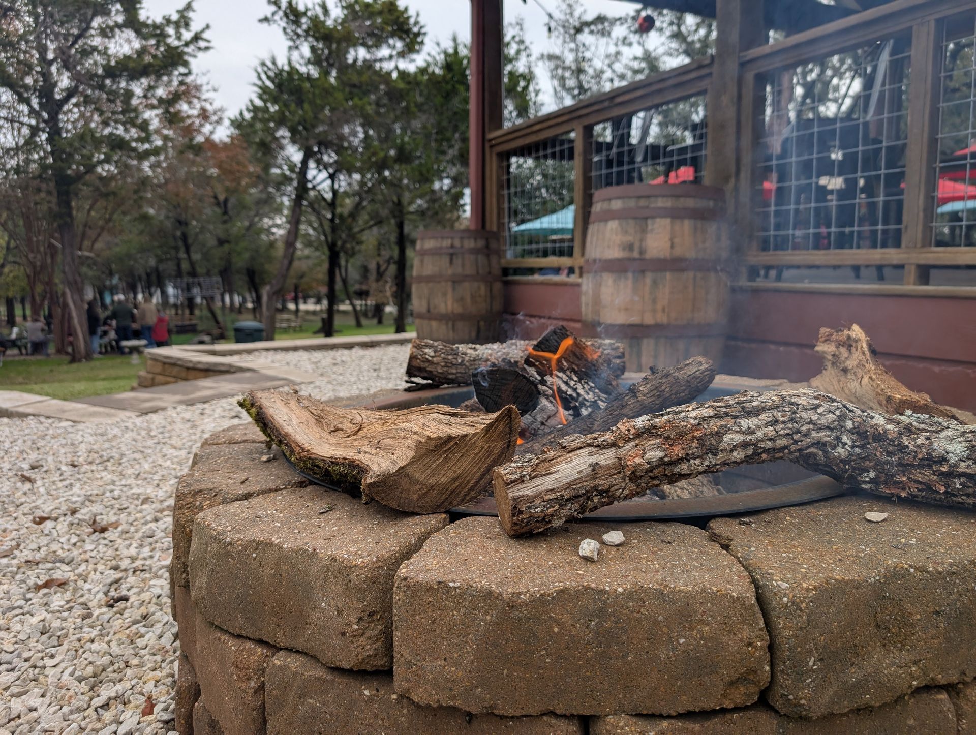 A brick fire pit with a small fire burning, next to wood barrels; outdoor setting.