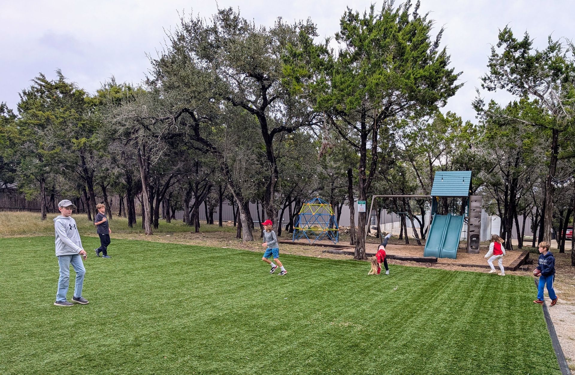 Children playing on a green turf field, a slide and trees in the background on a cloudy day.