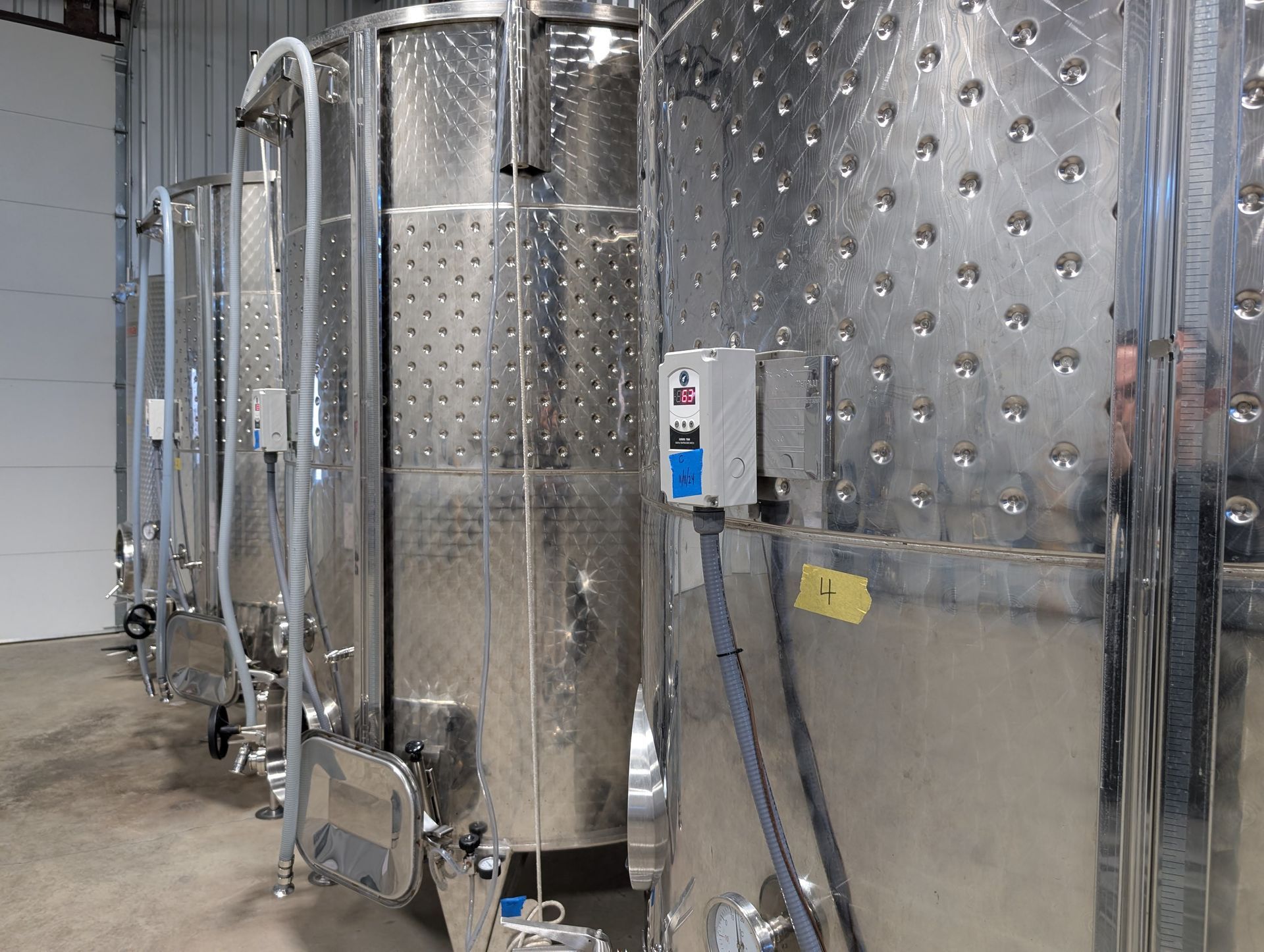 Row of stainless steel fermentation tanks in a winery. They have dimpled walls and various gauges and attachments.