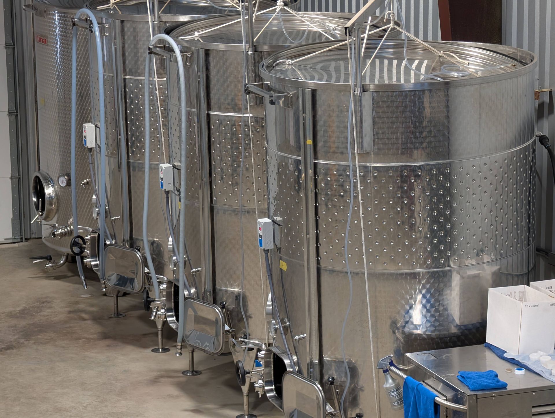 Stainless steel fermentation tanks in a winery, arranged in a row, with attached tubing.
