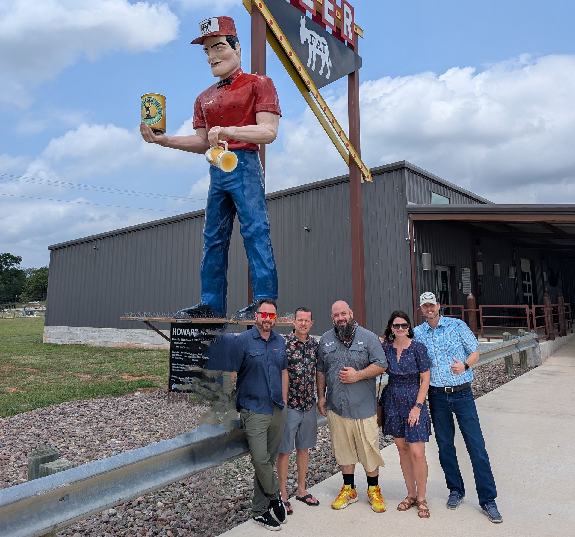 Group of five people posing by a giant statue of a man holding a beer can outside a building.
