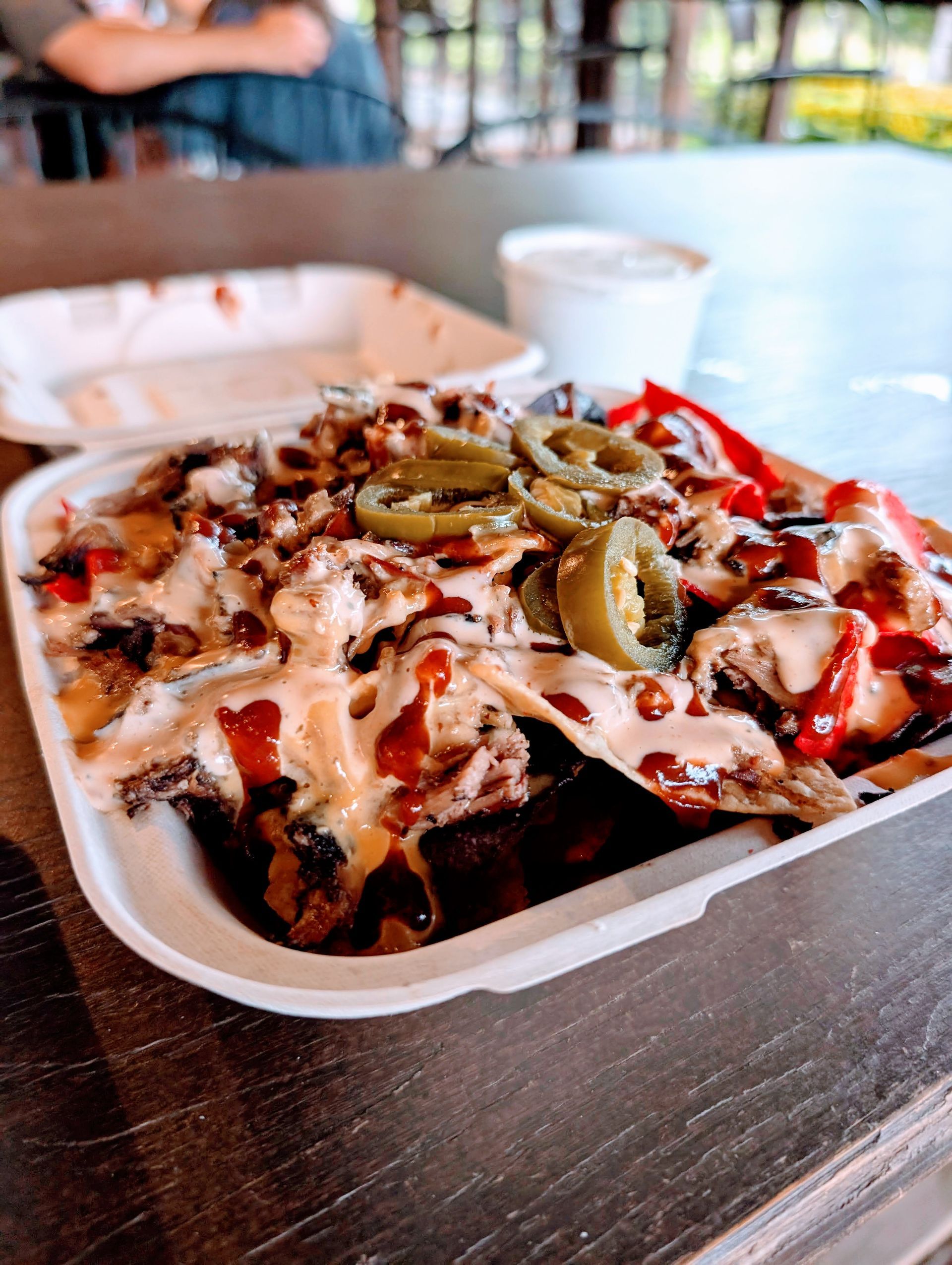 Nachos in a takeout container, topped with sauce, jalapenos, and meat.