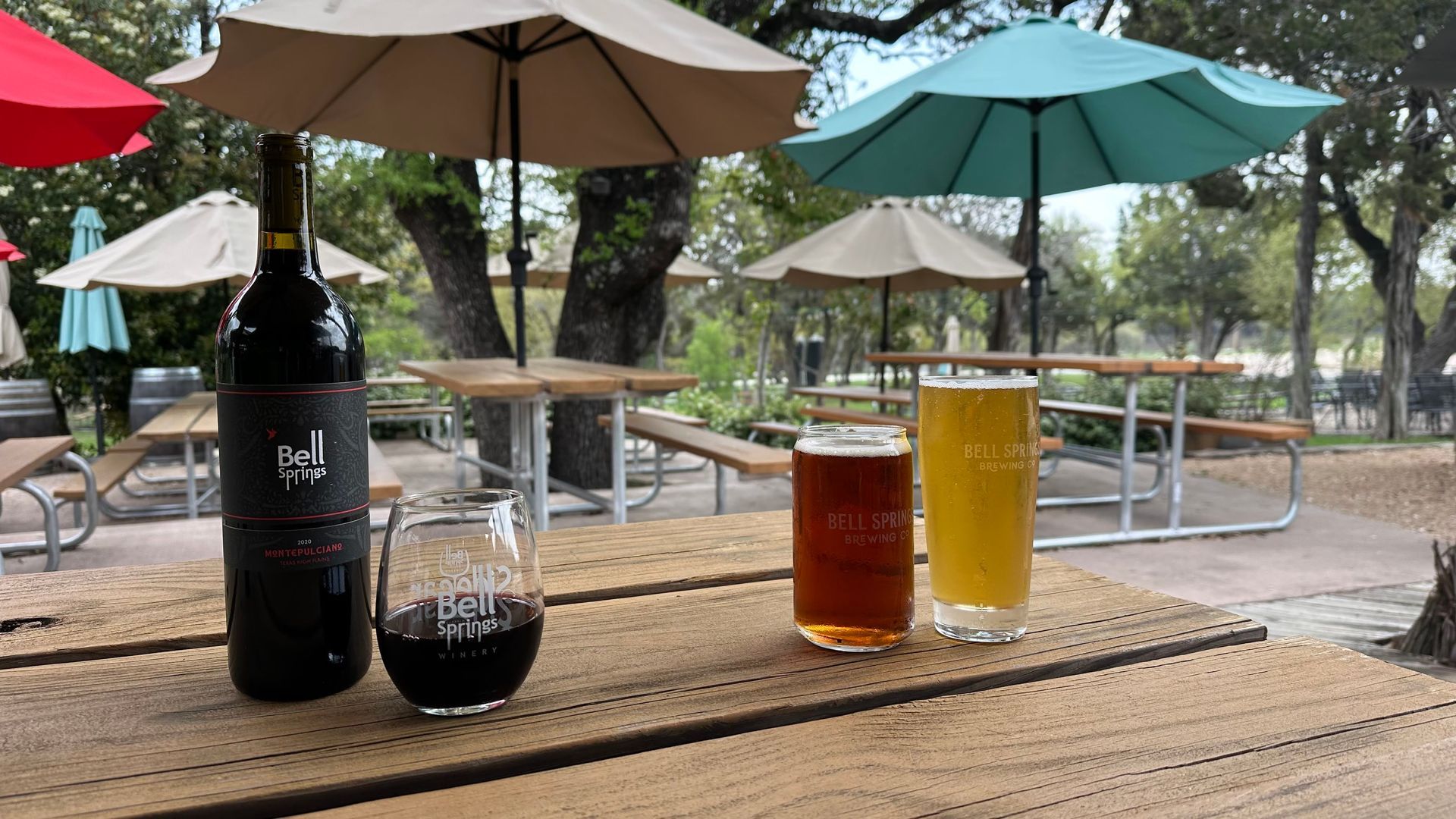 A bottle of wine and two glasses of beer are on a table under umbrellas.