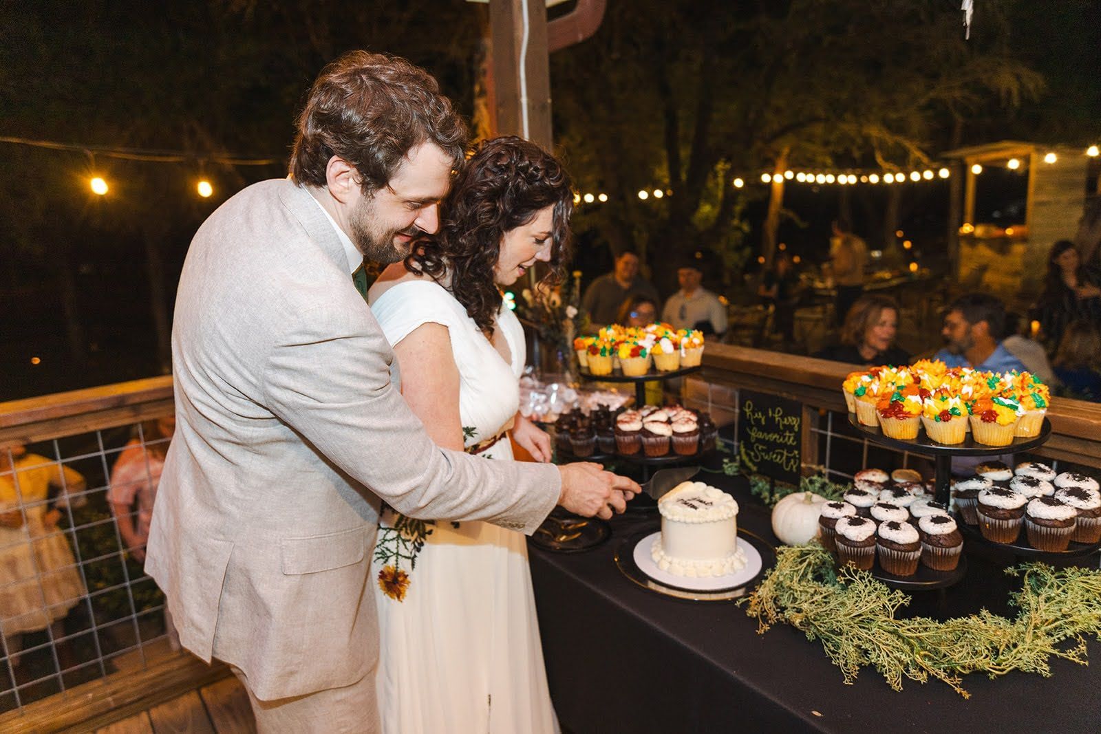Bride and groom cutting small cake at outdoor wedding, surrounded by cupcakes, guests visible.