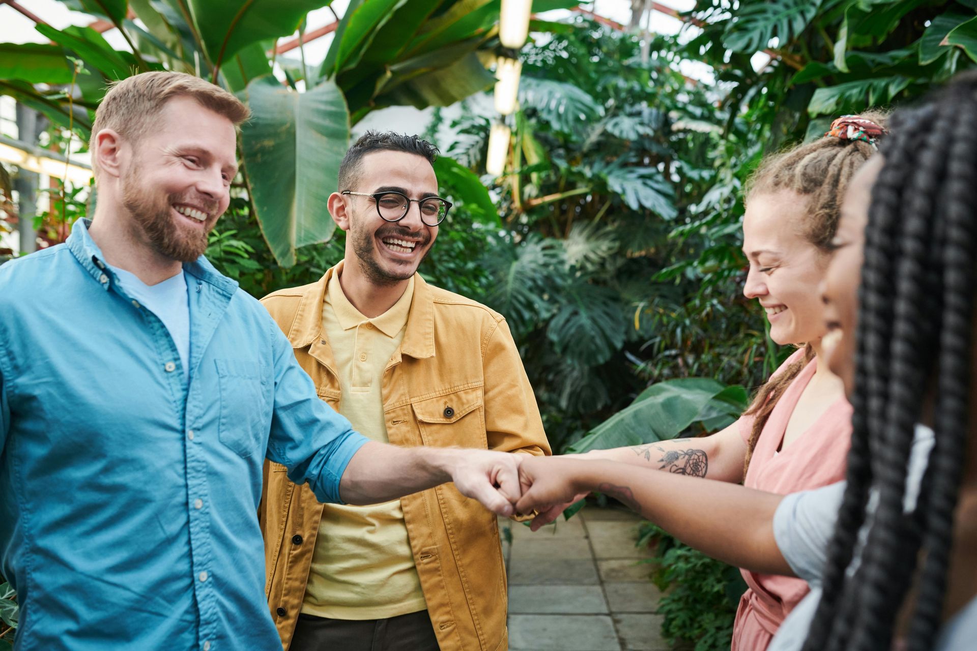 Group of friends fist-bumping, smiling, and laughing in a greenhouse with lush greenery.