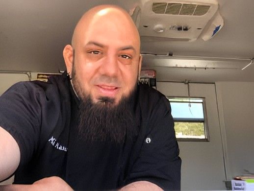 Bald man with a black beard in a food truck, wearing black chef's jacket, looking at camera, neutral expression.