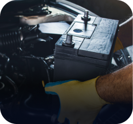 A close-up of a person in yellow gloves installing a black car battery into an engine bay | Ames Automotive STC