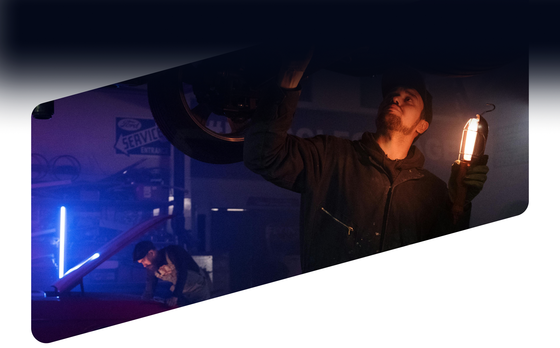 A mechanic illuminates the undercarriage of a car in a dimly lit garage while another person works in the blue-lit background | Ames Automotive STC
