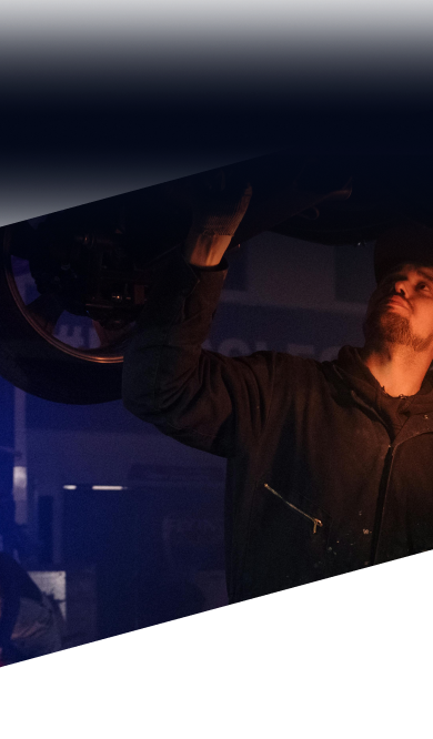 A technician in a dark shop inspects the underside of an elevated vehicle, illuminated by warm orange lighting | Ames Automotive STC