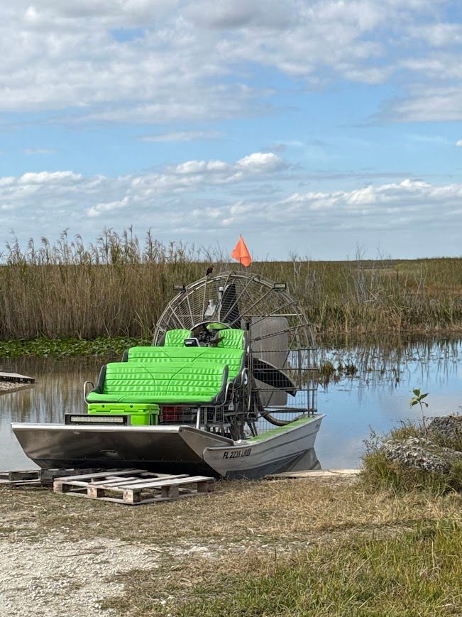 Airboat with green seats and propeller on water near marsh.