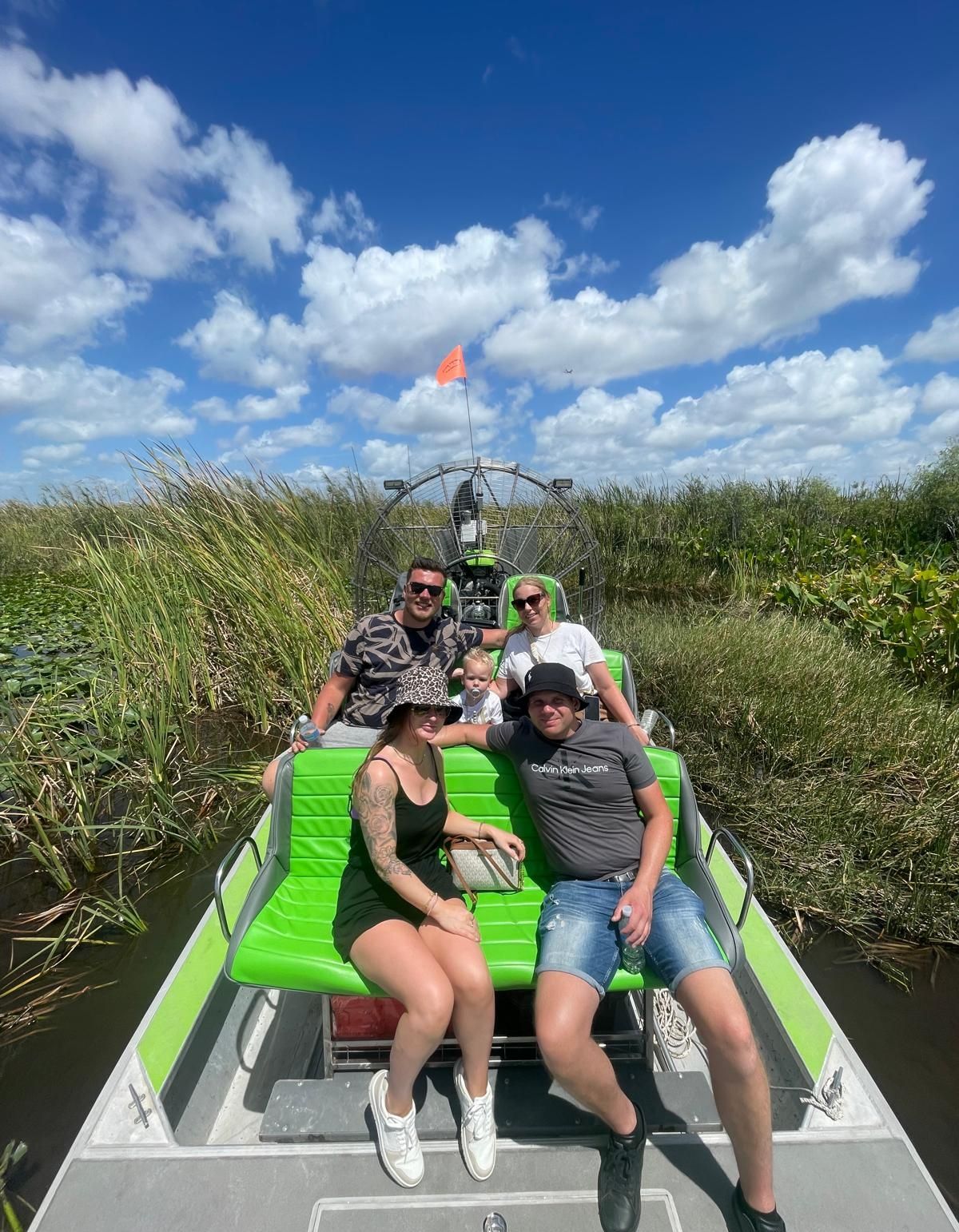 People on a neon green airboat in a swamp, sunny blue sky with clouds.