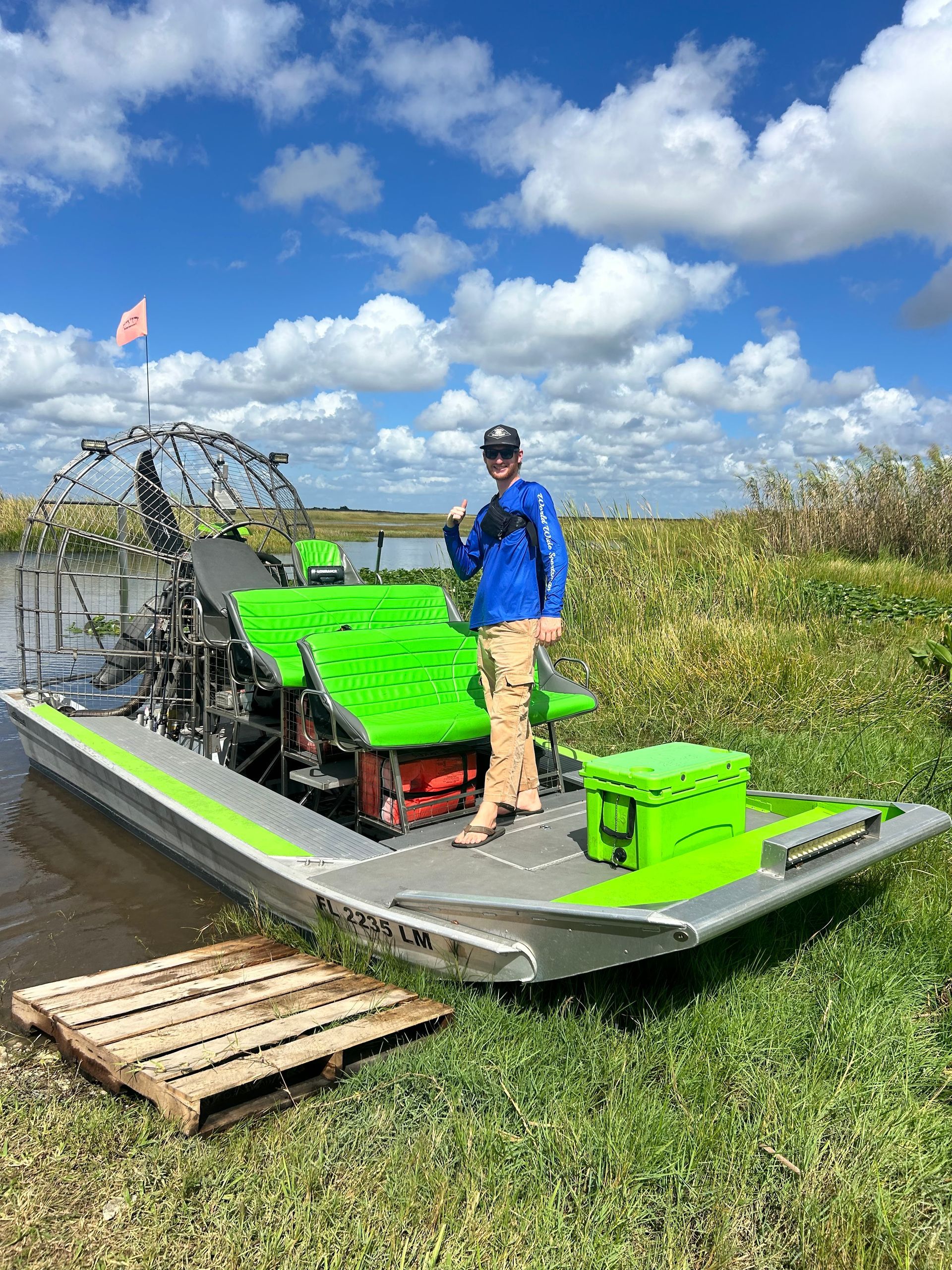 Person standing next to a lime green and silver airboat on a sunny day in a grassy marsh.