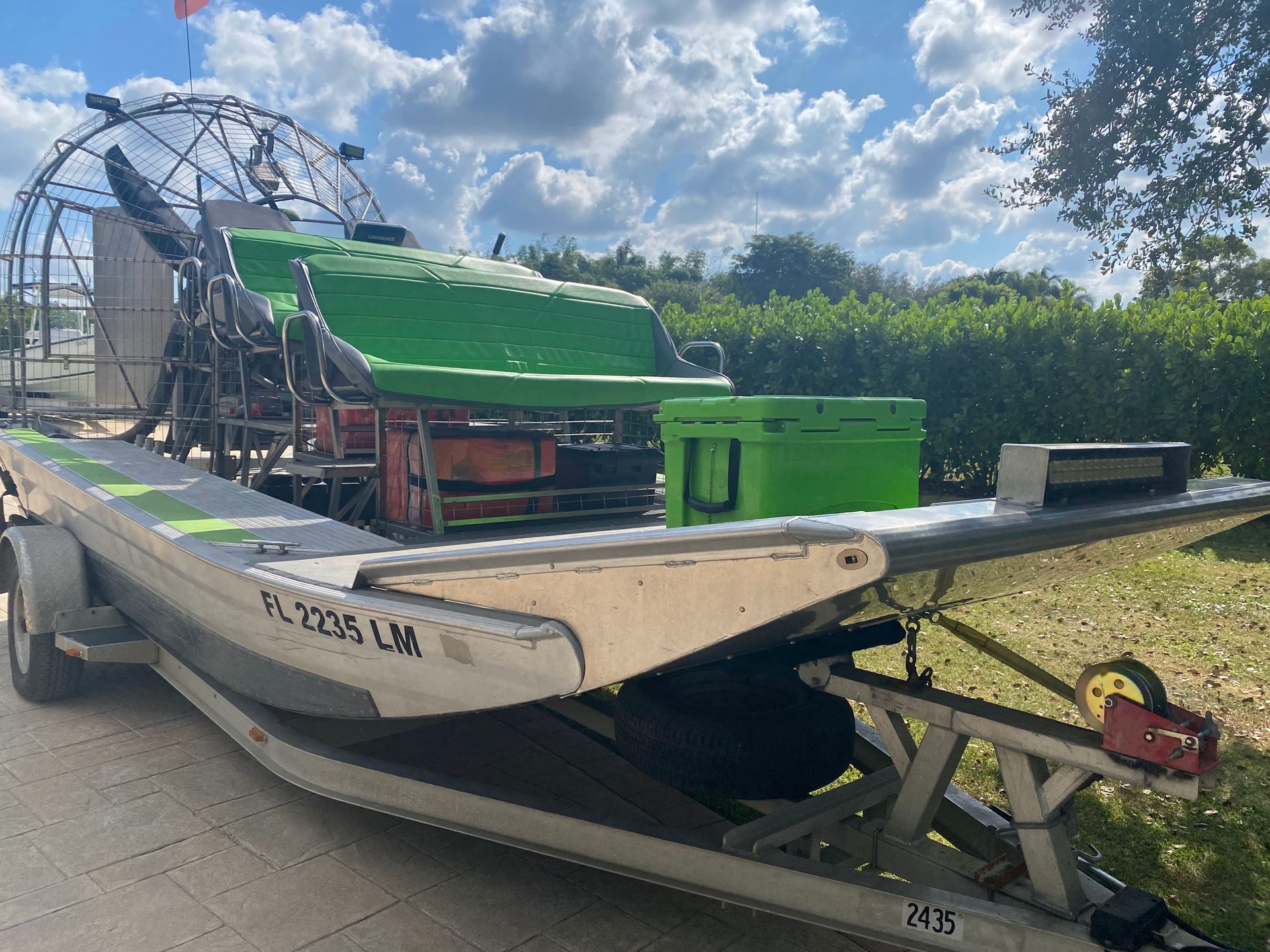 Airboat on trailer, green seats, cooler. Outdoor daytime with blue sky, and green hedge background.