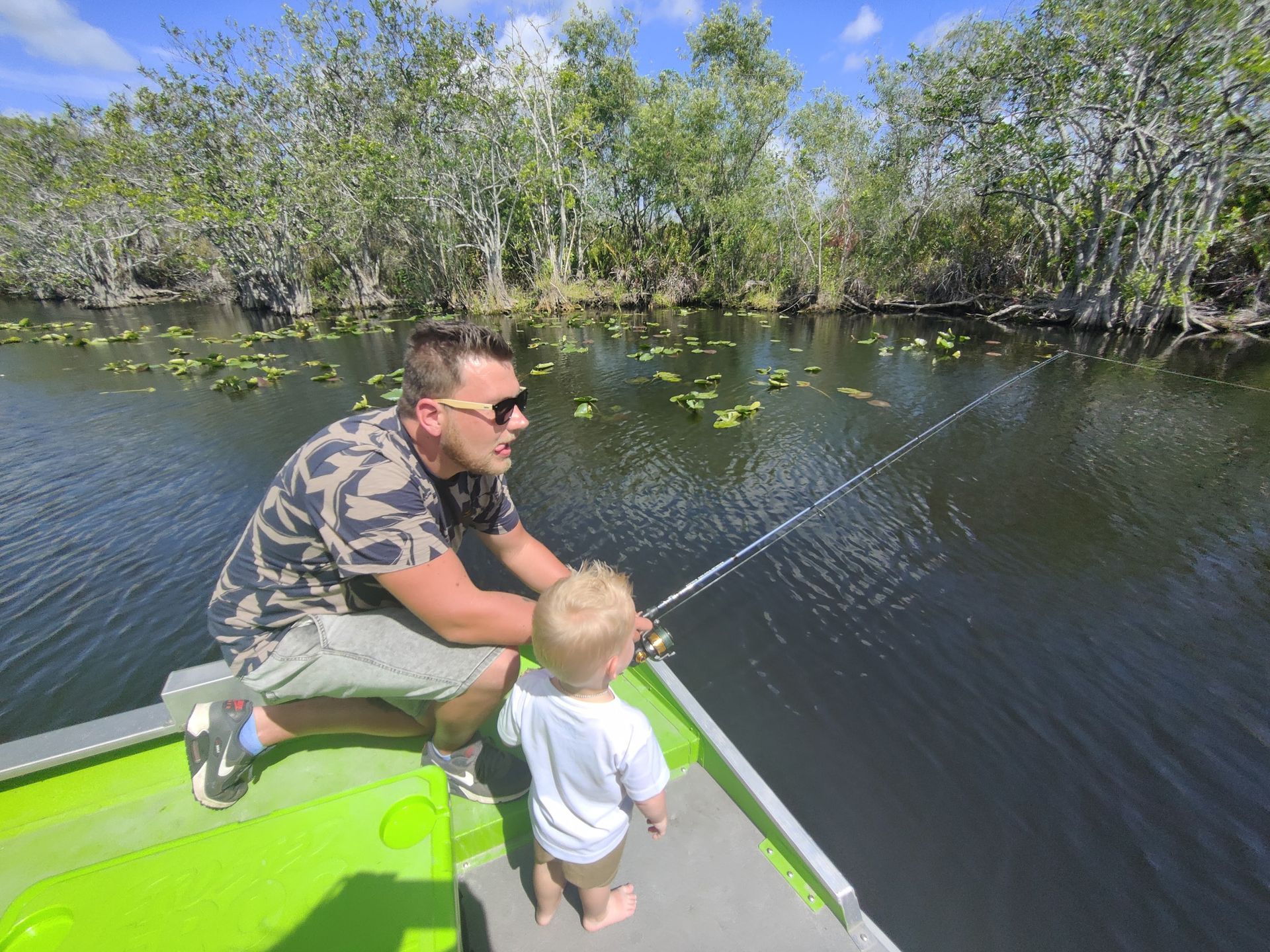 Man and toddler fishing from a boat on a dark waterway, surrounded by trees.