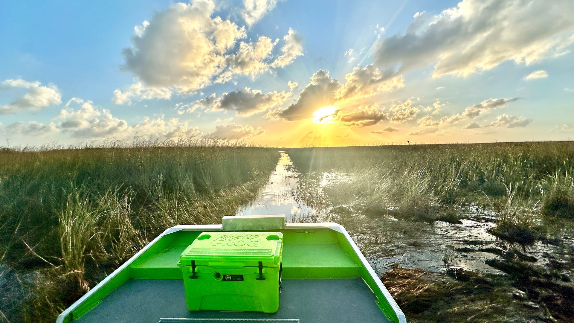Boat traveling through marsh at sunset, green cooler on deck, sunburst through clouds.